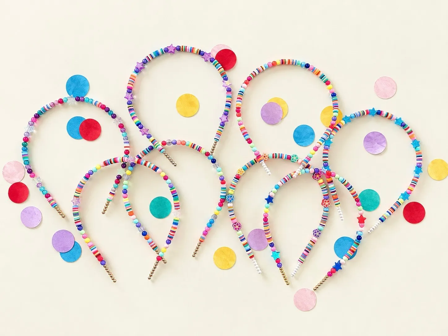 Nine headbands strung with rainbow clay disc beads, pearls, stars and flower charms, fanned out among scattered paper confetti.