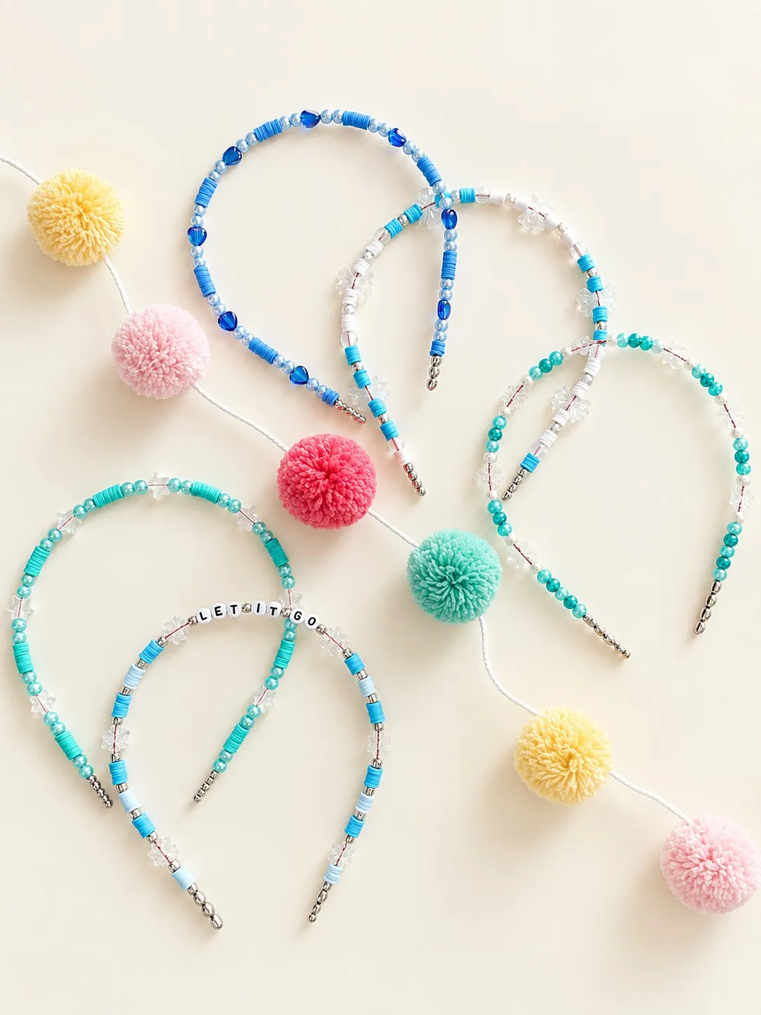 Five blue and aqua beaded headbands with clear snowflake beads, one spelling LET IT GO in letter beads, beside a pom-pom garland.