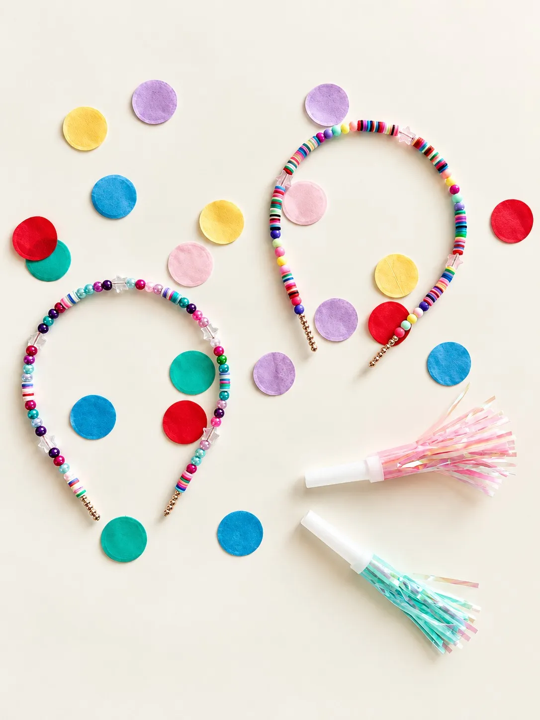 Two rainbow headbands of clay disc beads, pearls and clear stars, beside paper confetti and two iridescent party blowers.