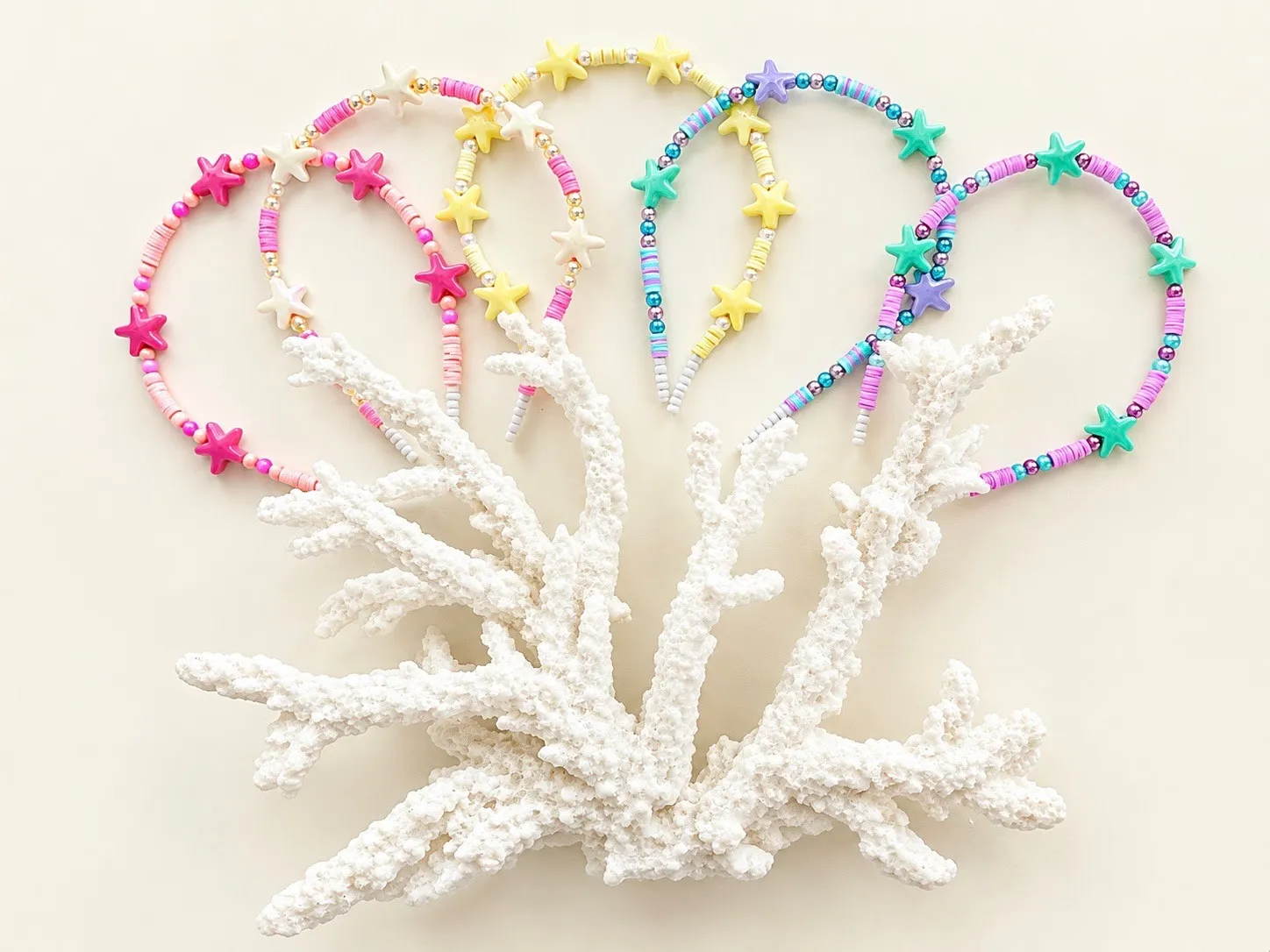 Five headbands with pink, yellow and teal starfish charms, fanned behind a branch of white coral.